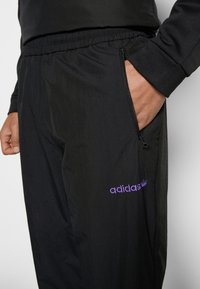 Svarta träningbyxor i lätt material, med elastisk midja, ficka med dragkedja och en lila "adidas"-logotyp.