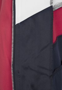 Primo piano di una cerniera di giacca con pannelli di tessuto rosso, bianco e blu navy e dettagli di cucitura visibili.