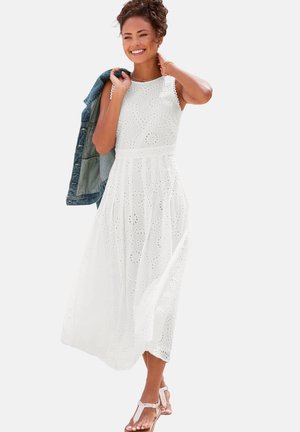 Weißes Midikleid aus Lochstickerei mit gewellten Abschlüssen und tailliertem Gürtel. Eine verblasste, blaue Jeansjacke ist über die Schulter gehängt.