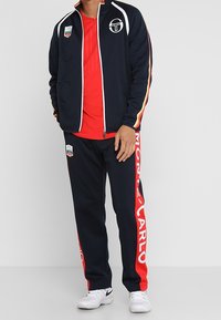 Laivastonsininen urheilupuku, jossa punaiset yksityiskohdat, vetoketjullinen takki, rintakehällä logotunnus ja "MONTE CARLO" -teksti housujen sivulla.