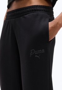 Κοντινό πλάνο ενός ατόμου που φοράει μαύρο παντελόνι φόρμας Puma με ελαστική μέση και χέρι στην πλαϊνή τσέπη, πάνω σε λευκό φόντο.