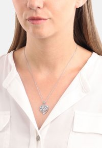 Söljfärgad hjärtformad hängsmycke med träddesign, hängande från en delikat kedja. Bärs över en vit blus, med fokus på halslinningen.