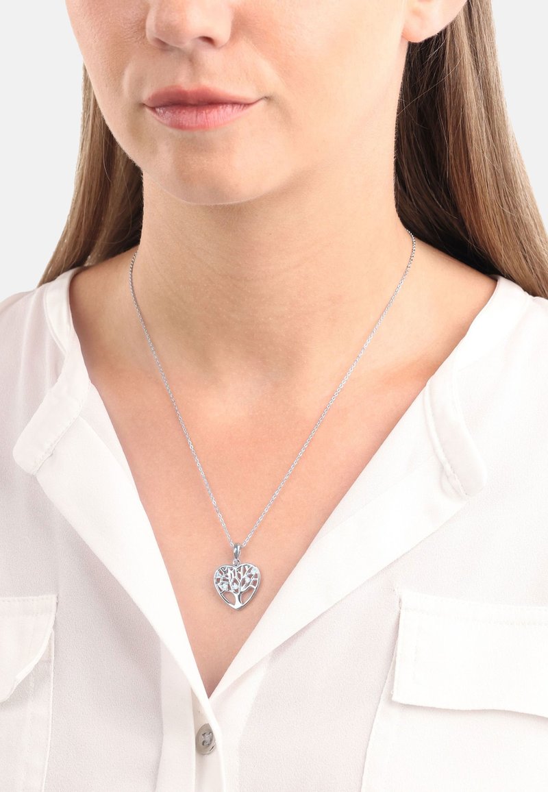 Söljfärgad hjärtformad hängsmycke med träddesign, hängande från en delikat kedja. Bärs över en vit blus, med fokus på halslinningen.