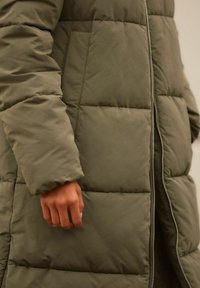 Olivovo zelená péřová bunda s prošívanou texturou, vybavená předním zipem, bočními kapsami a zatíženým manžetovým zakončením rukávu.