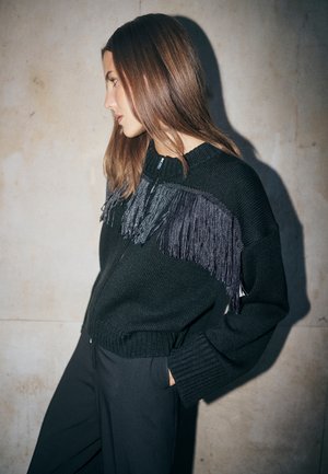 Svart stickad kofta med strukturerad design och fransdetalj i grå nyanser, med en avslappnad passform och dragkedja.