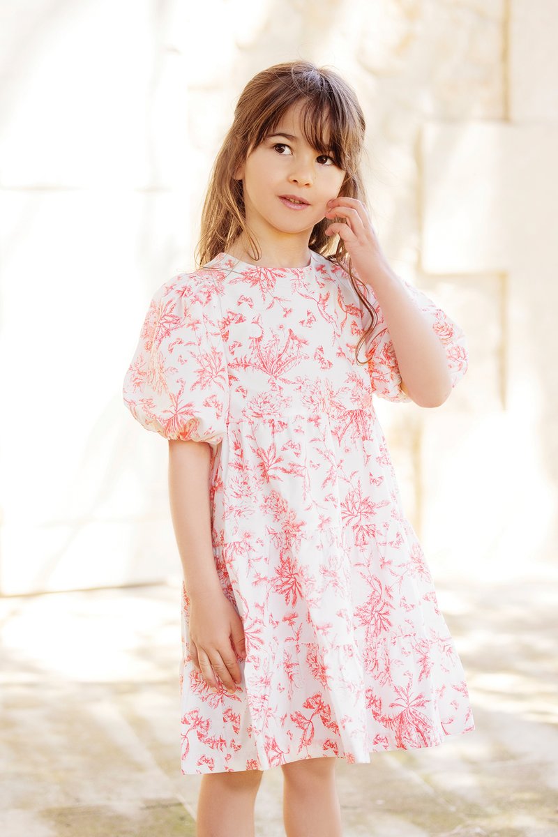 Vestido branco com padrões florais vermelhos, mangas curtas bufantes e decote redondo. Cai acima do joelho com uma saia franzida.