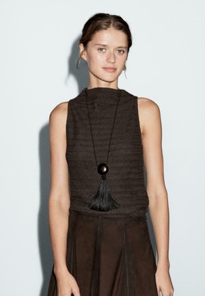 Brown textured sleeveless top with a high neck and a long black tassel pendant necklace. Model wears a dark brown textured skirt.