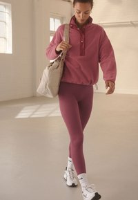 Polarowy pulower w jasnoróżowym kolorze z suwakiem do ćwiartki i detalem na guziki, zestawiony z dopasowanymi, prążkowanymi legginsami w ciemnoróżowym kolorze oraz białymi sneakersami.