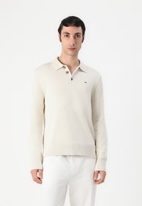 Beige kootud pullover polo kaelusega, kolme pruunist nupuga ning ribidega mansetid ja allääres. Väike logode detail vasakul rinnal.