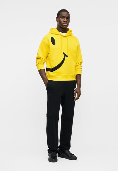 Άνδρας που φοράει κίτρινο φούτερ με κουκούλα με μεγάλο μαύρο σχέδιο χαμογελαστού προσώπου, μαύρο παντελόνι και μαύρα αθλητικά παπούτσια, στέκεται με τα χέρια στις τσέπες.