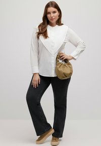 Camicia bianca a maniche lunghe con colletto piegato, abbinata a jeans neri e sneakers color beige. Tiene una piccola borsa a tracolla beige.