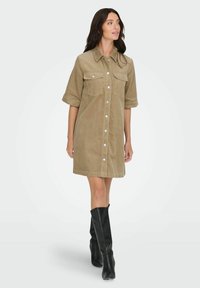 Haljina od korduroja u kaki boji s kratkim rukavima, kopčanjem na gumbe sprijeda, dva džepa na prsima i rubom do koljena, uparena s crnim čizmama koje sežu do koljena.