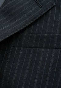 Ciężka tkanina garniturowa w odcieniach ciemnej wełny z delikatnym wzorem w prążki w odcieniach szarości i niebieskiego. Gładka tekstura z wyraźnie zarysowaną strukturą.