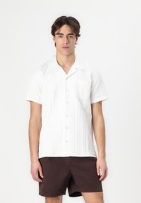 Camisa blanca de manga corta con un patrón texturizado, cierre de botones en la parte delantera y un bolsillo; combinada con pantalones cortos marrón oscuro.