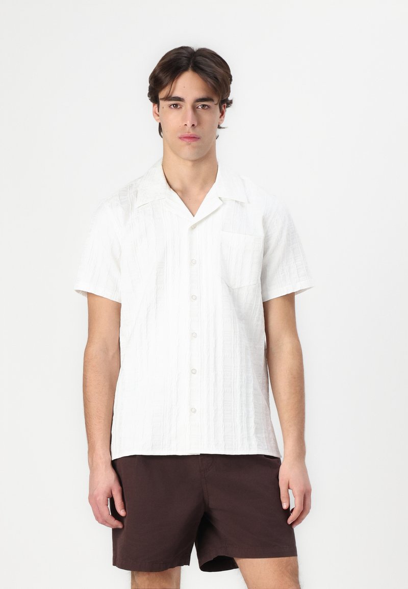 Camisa blanca de manga corta con un patrón texturizado, cierre de botones en la parte delantera y un bolsillo; combinada con pantalones cortos marrón oscuro.