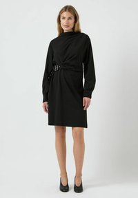 Černé šaty s dlouhým rukávem a upnutým siluetou, s navinutým designem a přezkou na pase. Hladká textura, po kolena.