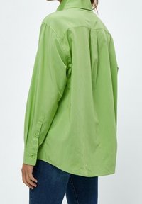 Camicia verde a maniche lunghe in tessuto morbido, con colletto alto, polsini con bottone e dettaglio plissettato sul retro. Abbinata a jeans blu.