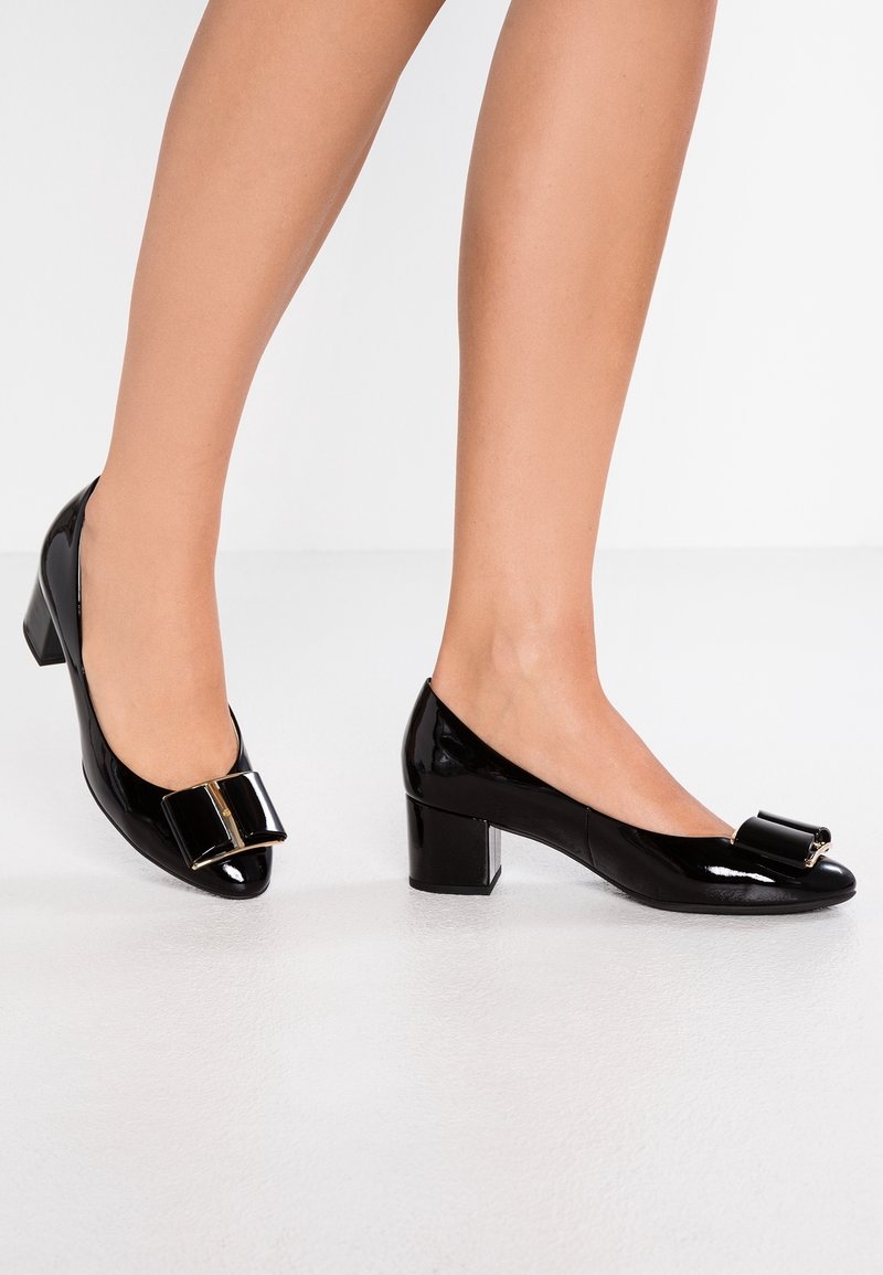 Ben som bär blanka svarta låga blockklackskor med stora matchande rosetter på ett vitt golv.