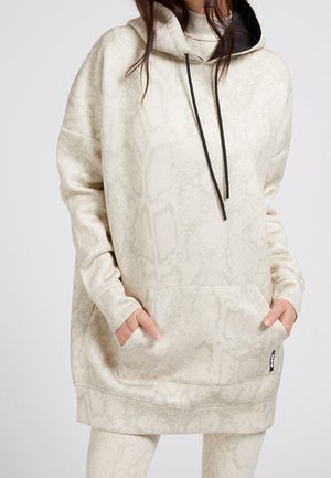 Femme portant un sweat à capuche beige imprimé serpent surdimensionné, avec poche avant et cordons noirs, mains dans les poches, visible des épaules aux cuisses.