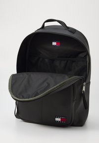Black backpack with a smooth, matte finish. Features a green zipper detail and mesh pocket inside. Logo patch visible on the front.