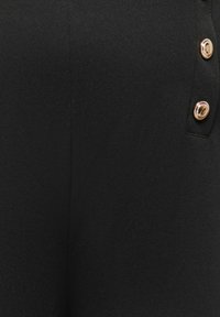 Sorte bukser i tekstureret stof med en åbning i siden med tre guldbelagte knapper og en glat finish.