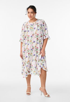 Kvinde iført en hvid knælængde kjole med blomsterprint, korte ærmer og flæsekant, kombineret med beige sandaler med høje hæle.