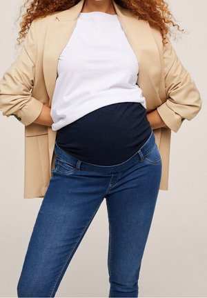 Kobieta w ciąży ubrana w beżową marynarkę, biały top i niebieskie jeansy ciążowe, z rękami opartymi na dolnej części pleców.