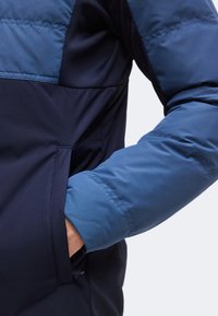 Personne portant une veste bicolore bleue avec la main droite insérée dans la poche latérale.