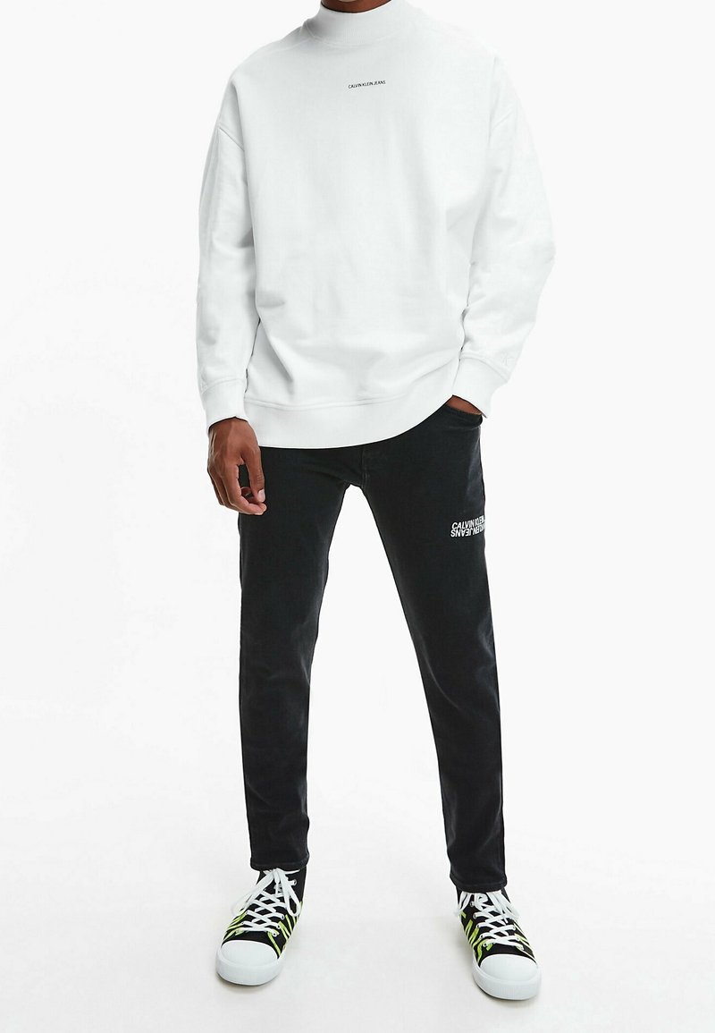 Sweatshirt en coton blanc avec des poignets côtelés, associé à un pantalon en denim noir arborant un logo de marque. Les chaussures comprennent des baskets noir et blanc avec des accents néon.