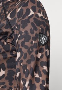 Leopárdmintás sportdzseki, sima, rugalmas anyagból, barna és fekete árnyalatokban, fekete EA7 logóval a kézelőn.