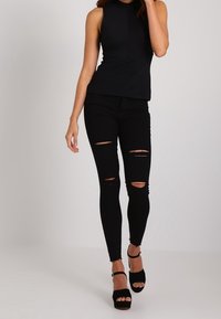 A woman in a sleeveless black top and ripped black jeans walks confidently in black high-heeled shoes against a neutral background.