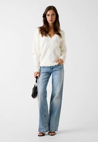 Pull en maille blanche et duveteuse avec un col en V, associé à un jean large bleu clair. Les accessoires comprennent un petit sac à main noir et des sandales noires.