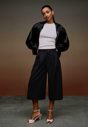 Nainen seisoo käsineen taskuissa, pukeutuneena mustaan nahkatakkiin, valkoiseen istuvaan paitaan, leveälahkeisiin tummanharmaisiin raidallisiin culottes-housuihin ja valkoisiin korkokenkäsandaaleihin.