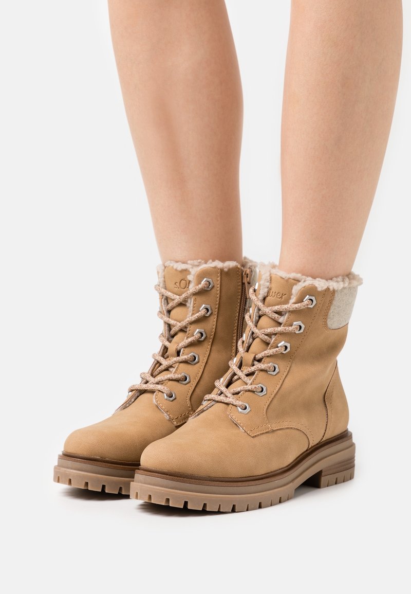 Botines de ante marrón con forro de pelo sintético, ojales de metal y diseño con cordones. Presentan una suela de goma resistente y suaves acentos de tela.