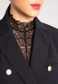 Czarny żakiet z wysokim koronkowym kołnierzem w misterny kwiatowy wzór, ozdobiony dwoma złotymi guzikami z przodu.