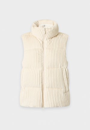 Jeune femme debout, main dans la poche, portant une veste matelassée crème, une chemise blanche à manches longues, un jean large bleu clair et des baskets blanches.