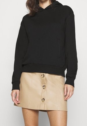 Sweat à capuche noir avec une poche kangourou, associé à une mini-jupe en cuir synthétique beige avec des boutons à l'avant et une texture lisse.