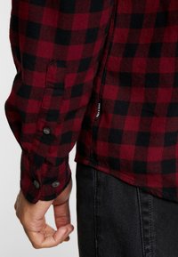 Rode en zwarte geblokte flanellen shirt met knoopmanchetten en zijsplitten. Label zichtbaar aan de zijkant. G gedragen over donkere jeans. Zachte textuur, normale pasvorm.