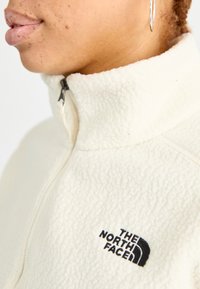 Giacca in pile di colore crema con colletto alto, caratterizzata da un logo ricamato nero di The North Face sul lato sinistro.