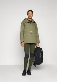 Olijfgroen jack met capuchon, voorzakken en verstelbare manchetten. Olijfgroene leggings met donkergrijze kniestukken, zwarte rugzak en zwarte schoenen.
