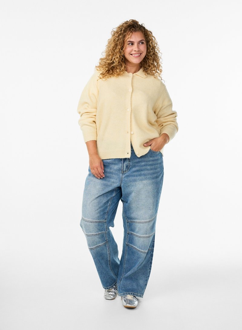 Mujer con cabello rizado que lleva un cárdigan crema, vaqueros azules holgados y zapatos plateados, de pie con una mano en el bolsillo y sonriendo.