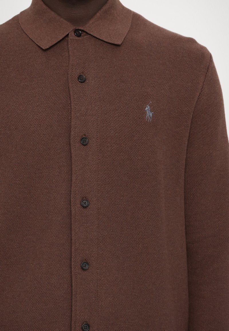 Braun gestricktes Polohemd mit strukturiertem Muster, ausgestattet mit einem geraden Kragen, schwarzen Knöpfen und einem kleinen, hellblauen Logo auf der linken Brust.