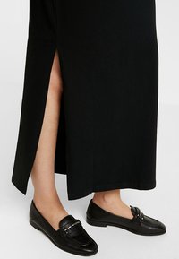 Abito lungo nero con spacco laterale, realizzato in tessuto morbido. Abbinato a mocassini neri con un dettaglio di catena argento sulla parte anteriore.