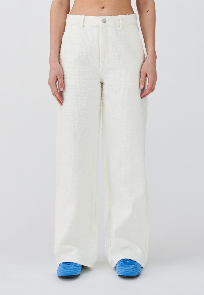 Pantaloni bianchi a gamba larga con texture, dotati di un bottone e zip frontali visibili, con tasche laterali, abbinati a scarpe da ginnastica blu.