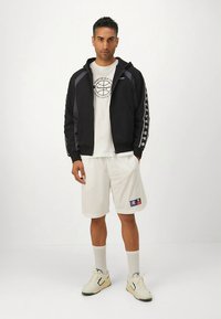 Casaco preto com fecho de correr, t-shirt branca com logótipo, calções brancos, ténis creme com detalhes verdes; apresenta riscas contrastantes e detalhes da marca.