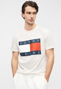 Φωτεινό μπεζ βαμβακερό t-shirt με ορθογώνιο γραφικό που περιλαμβάνει μπλοκ σε ναυτικό μπλε, λευκό και κόκκινο, και το κείμενο "TOMMY JEANS" σε ναυτικό μπλε.