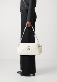 Borsa bianca trapuntata con un'accattivante decorazione di insetto dorato, tracolla a catena, forma rettangolare e superficie liscia, tenuta da una persona in abbigliamento nero.