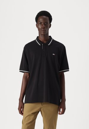 Jeune homme portant un polo noir à manches courtes avec des bordures blanches et un pantalon beige, se tenant devant un fond blanc uni.