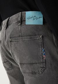 Grijze denim jeans met een blauw merklabel, met contrasterende stiksels in blauw en roze op de achterzak.