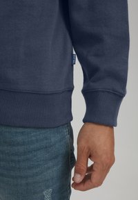 Felpa blu navale con polsini e orlo a costine, realizzata in morbido cotone misto. Presenta un piccolo etichetta sul lato sinistro vicino all'orlo.